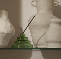 The Verde Glass Meso Incense Holder on a cream background with white pots behind it. 