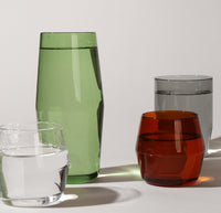 A 6 oz Clear and Amber Century Glass in front of a 16 oz Verde and 12 oz Gray Century Glass filled with water on a grayish background. 