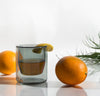 A 6 oz Gray Double Wall Glass filled with juice next to an orange on a cream background. 