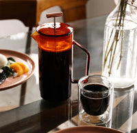 A Amber Glass French Press and Double Wall Glass filled with Coffee on a glass table.