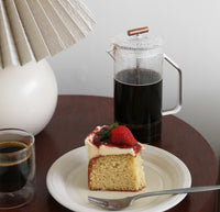 A Clear Glass French Press with coffee on a table with a slice of cake in front of it  on a gray background