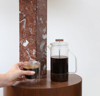 A Clear Glass French Press and a Clear Double Wall glass filled with coffee on a gray background.