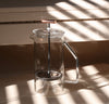 A Clear Glass French Press on a cream background with a shadow falling on it. 