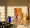 A Cobalt Glass French Press on a cream kitchen counter top with kitchen utensils next to it. 