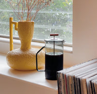 A Gray Glass French Press filled with coffee on a window sill with a yellow vase behind it. 