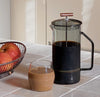 A Gray Glass French Press filled with black coffee and a Clear Century Glass filled with Milk coffee on a kitchen counter. 