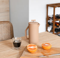 A Matte Sand Ceramic French Press with a Double Wall Glass filled with Coffee next to it on a wooden table.  
