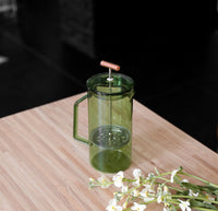 A Verde Glass French Press on a wooden table with flowers in the lower right corner.