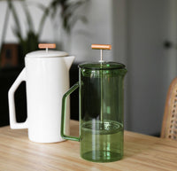 A Verde Glass French Press with a White Ceramic French Press behind it.