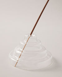A close up of the Clear Glass Meso Incense Holder with an incense stick, on a grayish background.