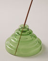 A close up of the Verde Glass Meso Incense Holder with an incense stick, on a grayish background.