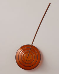 A top view of the Amber Glass Meso Incense Holder with an incense stick, on a grayish background.
