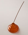The Amber Glass Meso Incense Holder with an incense stick, on a grayish background.