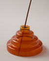A close up of the Amber Glass Meso Incense Holder with an incense stick, on a grayish background.
